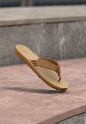 Hellbrauner Flip-Flop-Sandal mit gepolsterten Riemen, der über einer Steinoberfläche und vor einem grauen Stein-Hintergrund schwebt.
