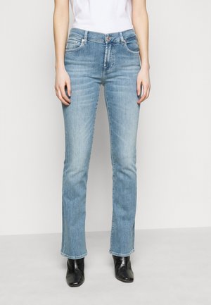 Person trägt hellblaue Jeans mit geradem Bein, weißes Oberteil und schwarze Stiefeletten, steht vor einem schlichten, hellen Hintergrund.