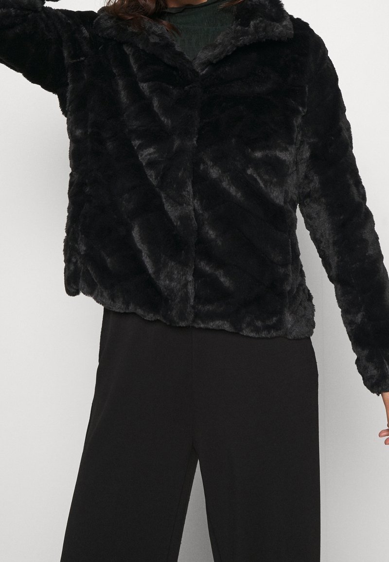 Personne portant une veste en fausse fourrure noire avec col et manches longues, associée à un pantalon noir sur un fond uni.