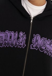 Felpa nera con zip, caratterizzata da un grande testo grafico viola che riporta le parole "DARK" e "NESS" in un carattere audace e stilizzato. Materiale in cotone.