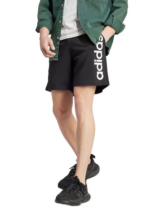 Zwarte shorts met het witte "adidas" logo aan de zijkant, gedragen met een groen overhemde en zwarte sportschoenen.
