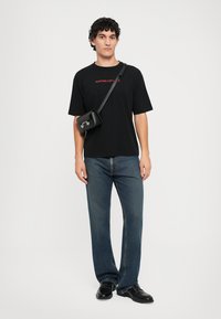 Svart T-skjorte med korte ermer og rød tekst, blå rette jeans, svart crossover-veske med sølvdetaljer, og svarte sko.