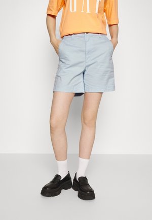 Person wearing light blue shorts, orange shirt with logo, white socks, and black chunky loafers, standing with hands in pockets.