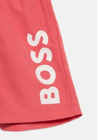 Κοντινό πλάνο υφάσματος κοραλλί-κόκκινου με λευκό κάθετο κείμενο "BOSS" τυπωμένο κοντά στην ραμμένη άκρη του τελειώματος.