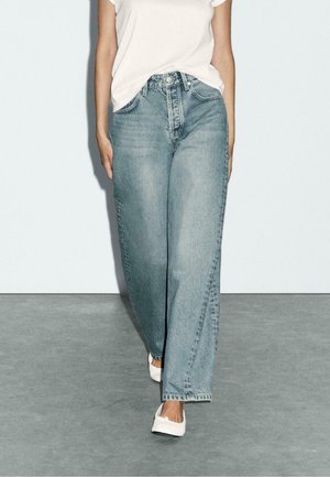 Helle blaue weit geschnittene Jeans aus Denim, mit hoher Taille und klassischem Fünf-Taschen-Design, kombiniert mit einem weißen T-Shirt und Slippern.