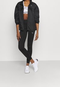Schwarze leichte Jacke mit Reißverschluss, gemusterter Cropped-Tops, schwarze Leggings und weiße Sneaker. Enthält Branding auf der Bekleidung und Kontrasttexturen.