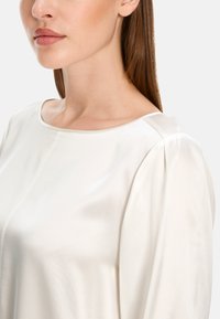 White satin blouse featuring a round neckline and subtle gathering at the shoulders. Smooth texture with a lightweight design.