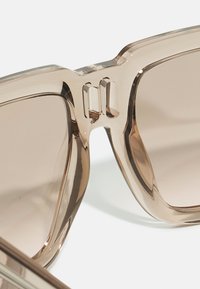 Ochelari de soare nuanța maro, cu rame din plastic transparent, voluminoase. Decupaje notabile la punte și o textură netedă, lucioasă.
