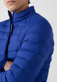 Man in een blauwe doorgestikte jas met de kraag omhoog, met gekruiste armen en een ronde logo-patch op de bovenste mouw.