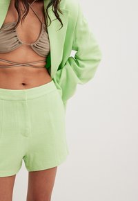 Hellgrüner strukturierter Blazer mit passendem kurzen Set; Unterbekleidung ist ein Taupe-farbener Bikini mit dünnen Trägern. Nahaufnahme des Outfits.