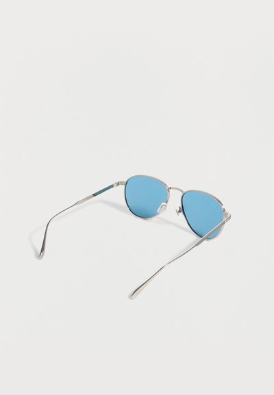 Γυαλιά ηλίου με ασημένιο μεταλλικό σκελετό, στρογγυλούς μπλε φακούς με απόχρωση, λεπτούς καμπυλωτούς βραχίονες και μαξιλαράκια μύτης, εμφανίζονται σε λευκό φόντο.