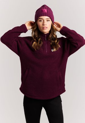 Jeune femme portant un pull polaire violet et un bonnet assorti, ajustant son bonnet avec les deux mains, debout devant un fond uni.