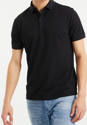 Mann trägt ein schlichtes schwarzes kurzärmeliges Poloshirt und hellblaue Jeans, steht mit entspannten Armen an den Seiten.