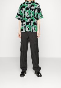 Blouse verte à manches courtes avec des motifs abstraits noirs et blancs ; associée à un pantalon cargo noir à rayures et des chaussures noires brillantes.