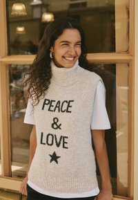 Gilet in lana beige con colletto alto, caratterizzato dalla scritta nera "PEACE & LOVE" e un design a stella. Indossato sopra una camicia bianca a maniche corte.
