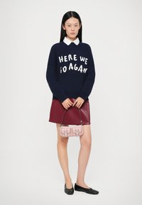 Maglione a maglia navy con la scritta bianca "ECCOCI ANCORA," gonna di pelle bordeaux, ballerine nere e una piccola borsa rosa con hardware in argento.