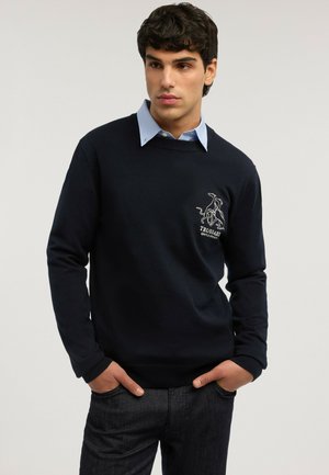 Junger Mann, der einen schwarzen Pullover mit einem Hundelogo und dem Text "Trussardi" über einem hellblauen Hemd mit Kragen trägt, die Hände in den Taschen, schaut zur Seite.