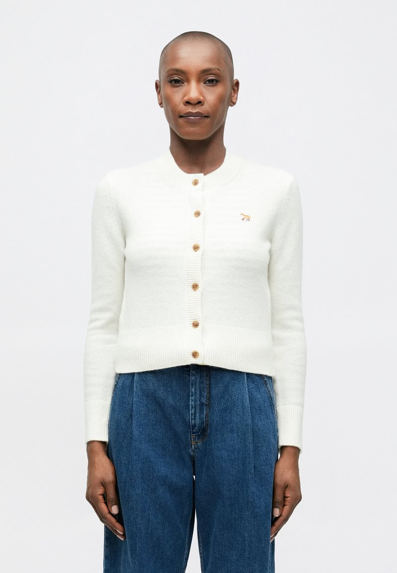 Cardigan blanco hecho de un tejido suave, con un diseño acanalado, cuello redondo y botones dorados. Tiene un pequeño detalle bordado.