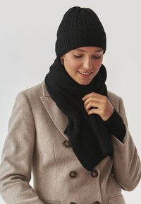 Un berretto lavorato a maglia nero con un motivo testurizzato, abbinato a una sciarpa nera. Il modello indossa un cappotto grigio chiaro doppiopetto.