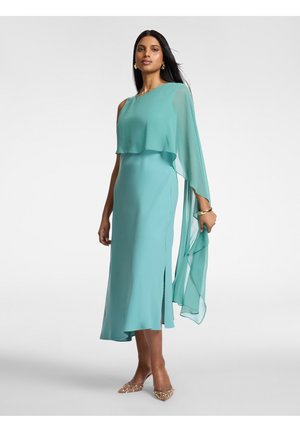 Vrouw draagt een teal midi-jurk met één schouder, met een doorzichtige overlay op één arm en gouden puntige hakken, staand tegen een effen achtergrond.