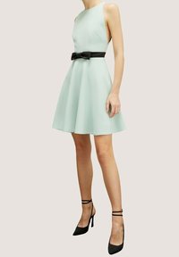Vihreä mintunvärinen hihaton mekko, jossa on levenevä helma, musta solmimisvyö vyötäröllä ja mustat korkokengät. Sujuva kankaan rakenne.