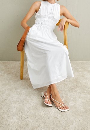 Vrouw in mouwloze witte jurk en witte sandalen met hakken en bandjes zittend op een houten stoel, met een tankleurige handtas aan de armleuning hangen.