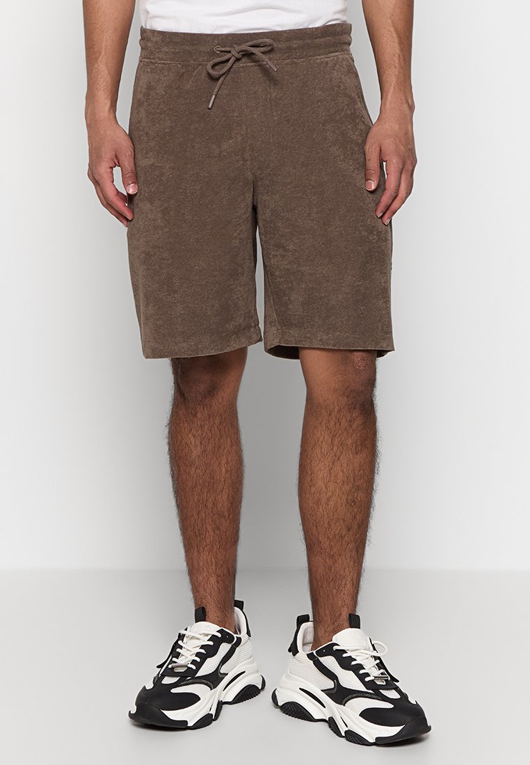 Braune Baumwollshorts mit elastischem Bund und Kordelzug. Weicher Stoff, knielang, kombiniert mit schwarz-weißen Turnschuhen.