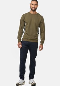 Sweatshirt vert olive avec un col rond, comportant des panneaux latéraux blancs avec du texte noir. Tissu en mélange de coton doux. Manches longues et poignets côtelés.