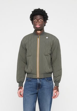 Veste bomber matelassée en vert olive avec un col montant, une fermeture éclair jaune et bleue, et des poignets élastiques, associée à un jeanm bleu.