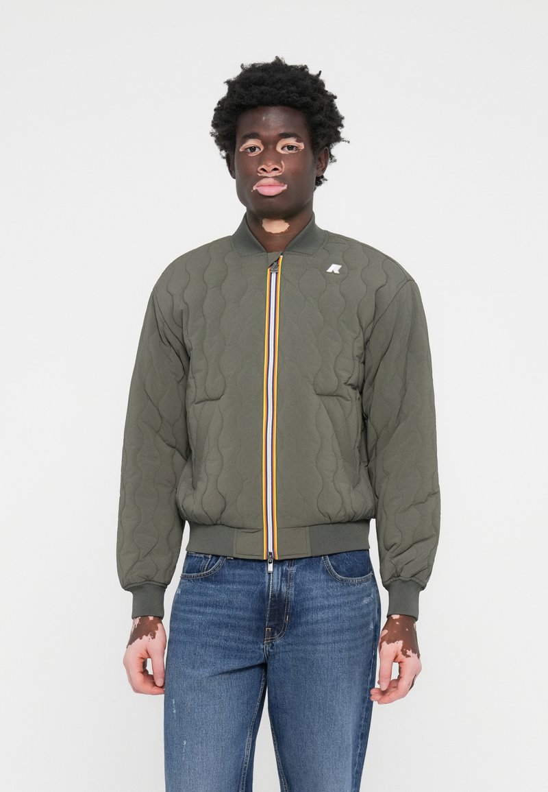 Olajzöld steppelt bomber dzseki álló gallérral, sárga és kék cipzárral, és elasztikus mandzsettákkal, kék farmerral párosítva.