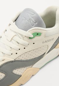 Lacoste sneaker featuring cream leather and grey accents, with perforated details, flat laces, and a green eyelet near the lacing.