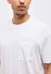 Άνδρας που φοράει απλό λευκό μπλουζάκι με στρογγυλή λαιμόκοψη και τσέπη στο στήθος, εμφανιζόμενος από τον λαιμό μέχρι τη μέση, με φόντο απλό λευκό.