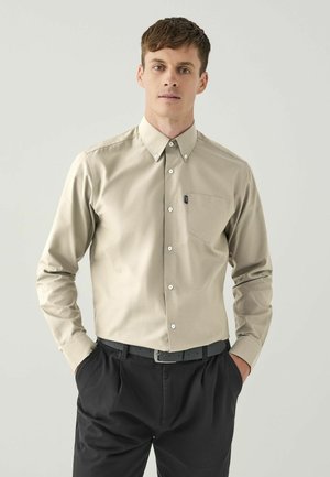 Beige Langarmhemd mit Kragen, einer Brusttasche und weißen Knöpfen, getragen mit schwarzen Hosen. Glatte Textur.