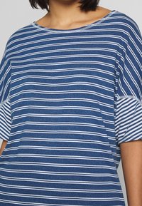 Camisa de manga corta a rayas azul y blanca, fabricada en un tejido suave. Presenta un escote ancho y un corte relajado con una textura sutil.