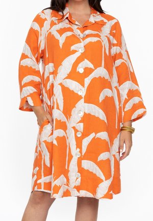 Luźna pomarańczowa sukienka z białymi wzorami liści palmowych, zapinana z przodu na guziki, z rękawami do łokci, bocznymi kieszeniami oraz złotymi bransoletkami na prawej ręce.