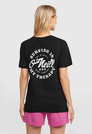 Čierne bavlnené tričko s veľkým bielym grafickým motívom na chrbte s nápisom "Surfovanie je moja terapia" a motívmi palmových stromov. Nosené s ružovými šortkami.