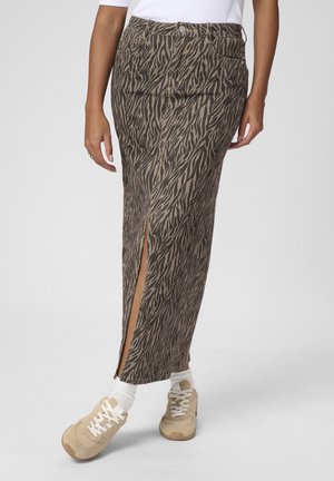 Femme portant une jupe longue marron à taille haute avec motif et fente à l'avant, associée à des chaussettes blanches, des baskets beiges et un top blanc.