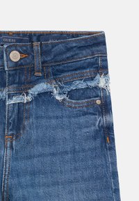 Calças de ganga em azul escuro com bainha desfiada, costura cobre e bolso frontal. Apresenta um botão de metal com a marca e um corte limpo.