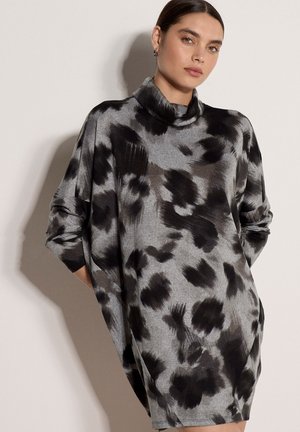 Femme aux cheveux plaqués en arrière porte une robe grise ample avec des motifs abstraits noirs et un col haut, posant devant un fond clair uni.