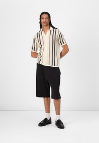 Kurzarm-Hemd mit Knöpfen in Creme mit schwarzen, orangen und grünen vertikalen Streifen. Kombiniert mit schwarzen Shorts und schwarzen Slippern.