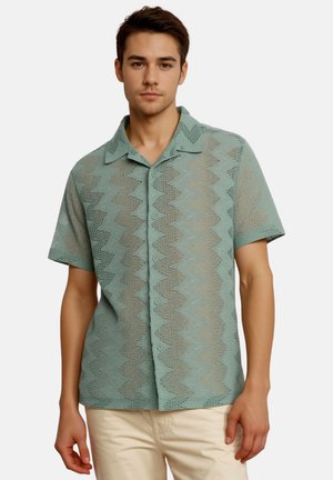 Młody mężczyzna w jasnozielonej, krótkorękawowej koszuli na guziki z zygzakowatym wzorem, w połączeniu z beżowymi spodniami.