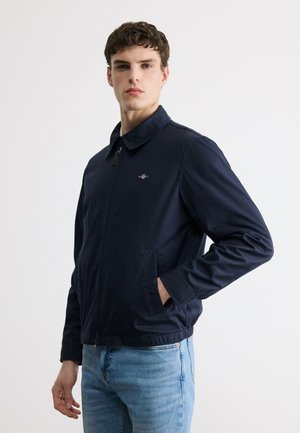 Ung mand iført en navyblå lynlåstrøje med krave og lyseblå jeans, stående mod en ensfarvet hvid baggrund.