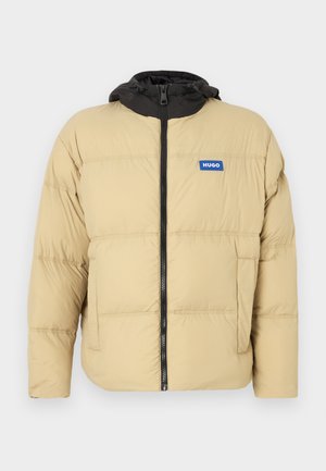 Béžová prešívaná bunda s čiernou kapucňou, predným zipsom a logom. Má prešívanú textúru a dve bočné vrecká.