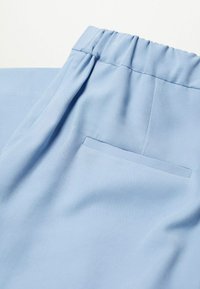 Pantalon bleu clair avec une ceinture élastique, une texture lisse et une petite poche arrière. Le tissu présente une légère apparence côtelée.