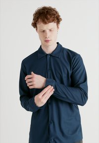 Dark blue button-up shirt with a textured ribbed fabric, featuring a collar and long sleeves. The model adjusts the cuff with one hand.