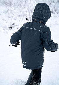 Črna priredjena zimska bunda z odebeljenim polnilom in kapuco, z belo poudarjeno črto in zlatim okovjem, ki jo nosi otrok v zasneženem okolju.