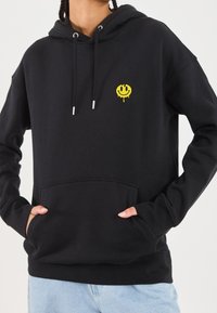 Sweat à capuche noir en coton avec une poche avant, des cordons de serrage réglables, et un logo de visage souriant jaune brodé sur la poitrine.