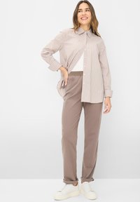 Camicia a maniche lunghe a righe marrone chiaro, abbinata a pantaloni tapered color taupe chiaro e sneakers bianche, su uno sfondo neutro.