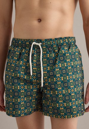 Primo piano di una persona che indossa pantaloncini da bagno con fantasia, con vita elastica e coulisse bianca, caratterizzati da forme geometriche in verde, giallo e rosso.