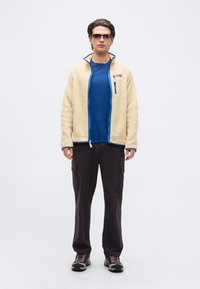 Άνδρας που φοράει μπεζ φλις τζάκετ, μπλε πουκάμισο, μαύρο μπλουζάκι τύπου cargo, παπούτσια πεζοπορίας και γυαλιά ηλίου, στέκεται μπροστά σε απλό φωτεινό φόντο.